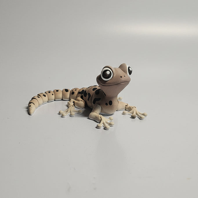 A small decorative lizard figurine with large cartoon eyes, spotted tan and cream coloring, and tiny black feet, placed on a white surface against a gray brick wall.